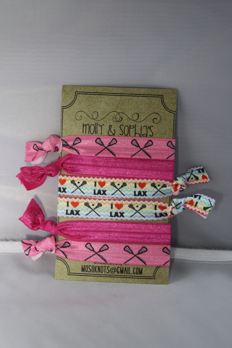 May include: A set of six pink and white hair ties with a lacrosse stick and heart design. The hair ties are packaged on a brown card with the text "molly & sophia's" and "mosoknots@gmail.com".