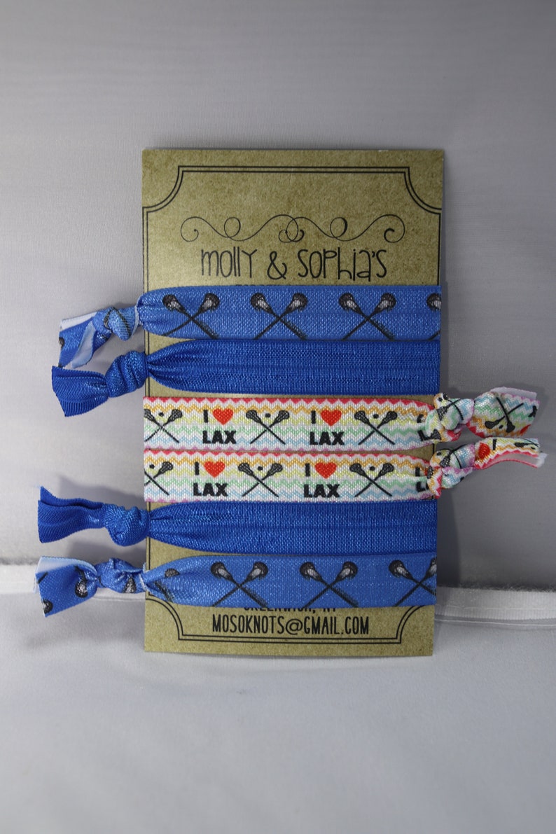 May include: A set of six blue and white lacrosse-themed hair ties. The hair ties are packaged on a brown card with the text "molly & sophia's" and "mosoknots@gmail.com".
