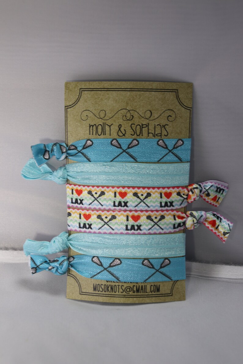 May include: A set of six blue and white lacrosse-themed hair ties with the text "I love LAX" on two of the ties. The hair ties are packaged on a brown card with the text "molly & sophia's" and "MOSOKNOTS@GMAIL.COM".