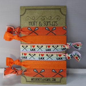 May include: A set of six orange and white elastic hair ties with lacrosse stick and heart designs. The hair ties are displayed on a brown card with the text "molly & sophia's" and "mosoknots@gmail.com".