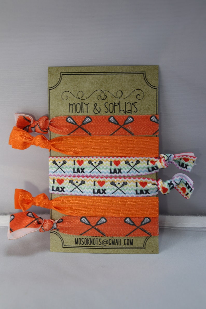 May include: A set of six orange and white elastic hair ties with lacrosse stick and heart designs. The hair ties are displayed on a brown card with the text "molly & sophia's" and "mosoknots@gmail.com".