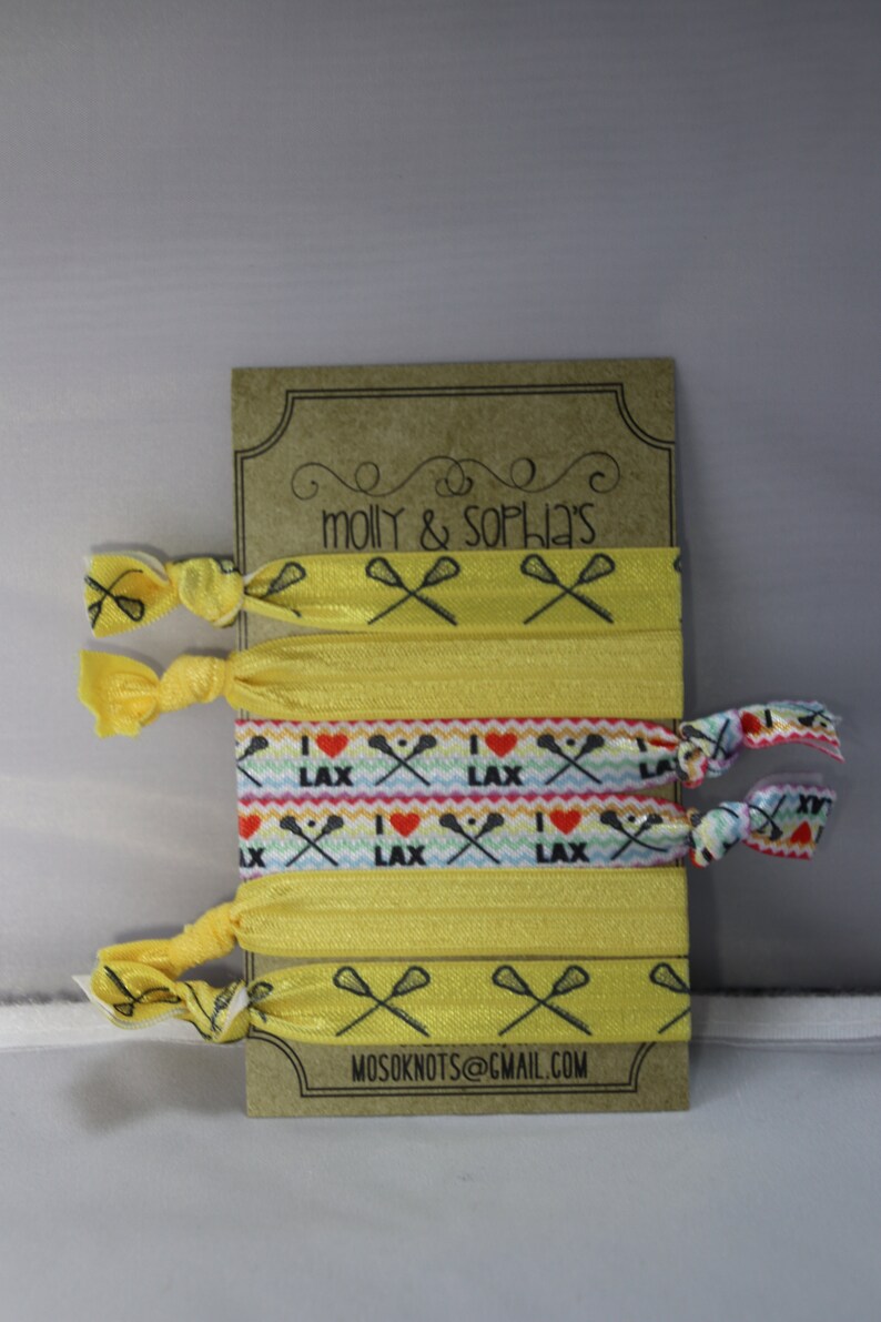 May include: A set of six yellow hair ties with lacrosse stick and heart designs. The hair ties are packaged on a brown card with the text "molly & sophia's" and "mosoknots@gmail.com".