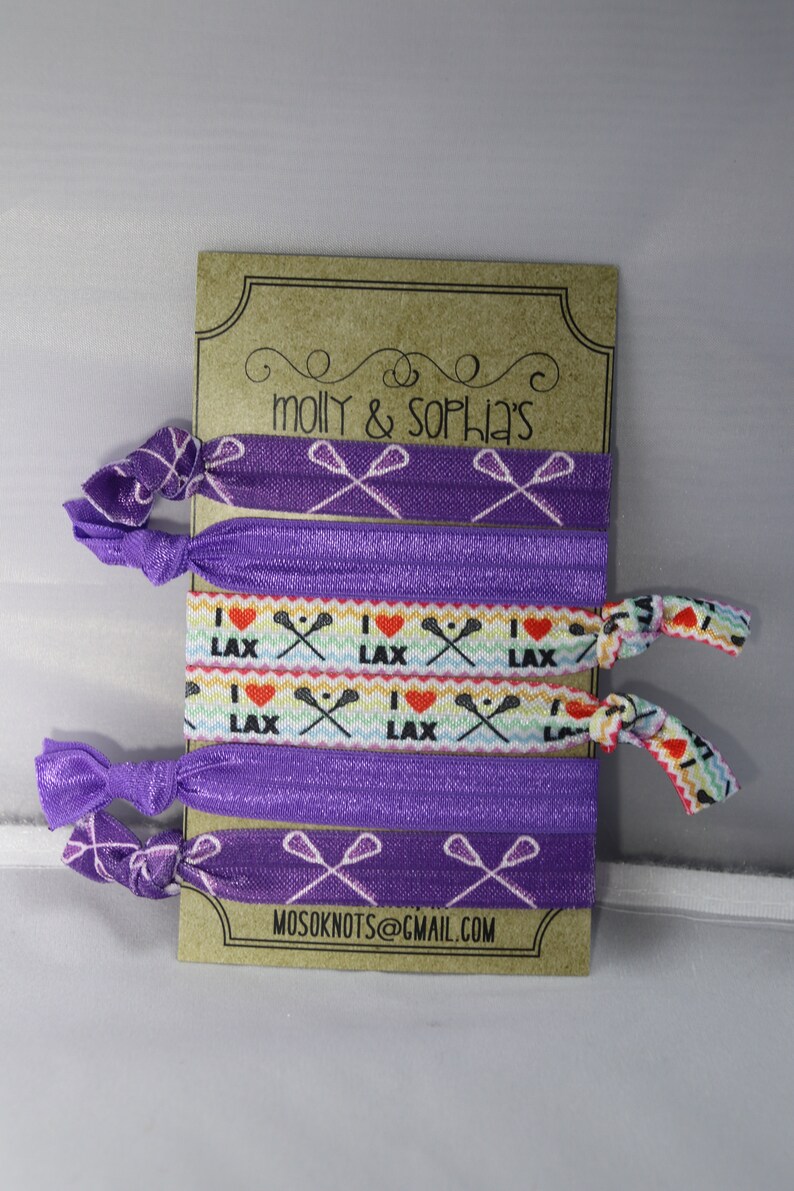 May include: Set of six purple elastic hair ties with lacrosse stick designs. Two hair ties feature a white lacrosse stick design on a purple background. Two hair ties feature a rainbow design with the words "I love LAX" and a white lacrosse stick design. Two hair ties feature a white lacrosse stick design on a purple background with white polka dots. The card reads "molly & sophia's" and "mosoknots@gmail.com".