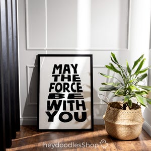 May include: Black and white framed print with the text "May the Force be with you" in a stylized font. The print is hanging on a white wall with a potted plant in a woven basket in the foreground.
