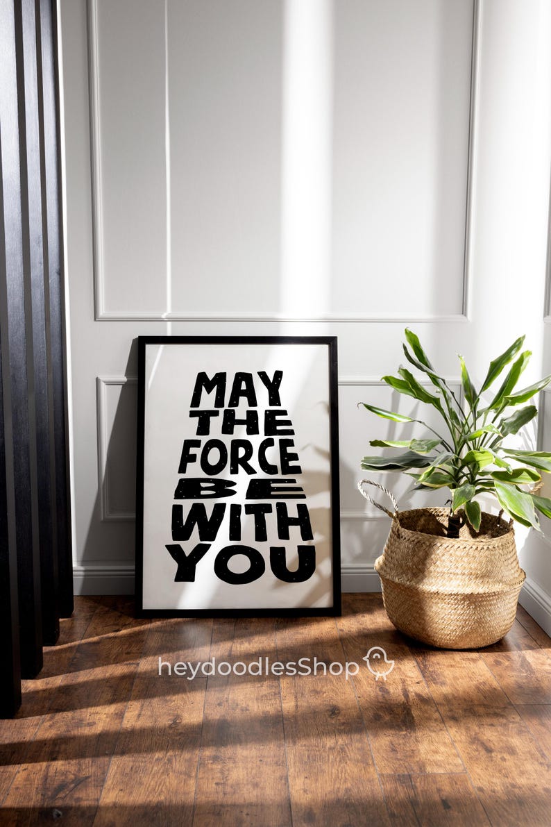 May include: Black and white framed print with the text "May the Force be with you" in a stylized font. The print is hanging on a white wall with a potted plant in a woven basket in the foreground.
