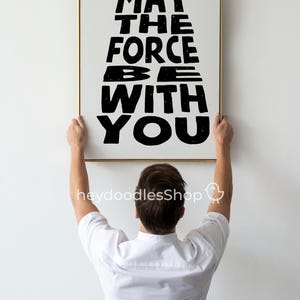 May include: A person hangs a framed print on a white wall. The print features the text "May the Force Be With You" in black lettering with a starry sky background.