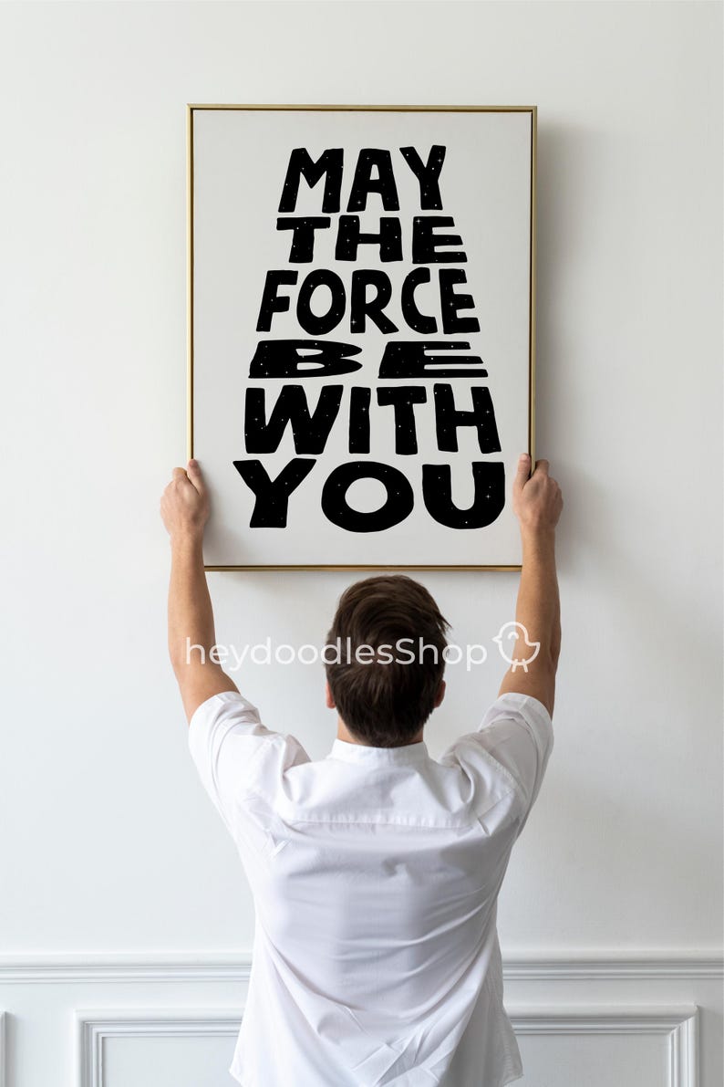 May include: A person hangs a framed print on a white wall. The print features the text "May the Force Be With You" in black lettering with a starry sky background.