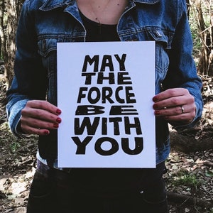 May include: A white poster with black text that reads "May the Force Be With You". The text is in a stylized font with a star-like pattern.