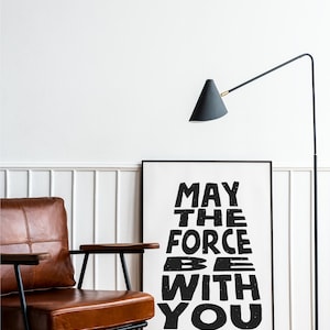 May include: Black and white typography print with the text "May the Force be with you" in a distressed font style. The print is framed and hanging on a white wall.