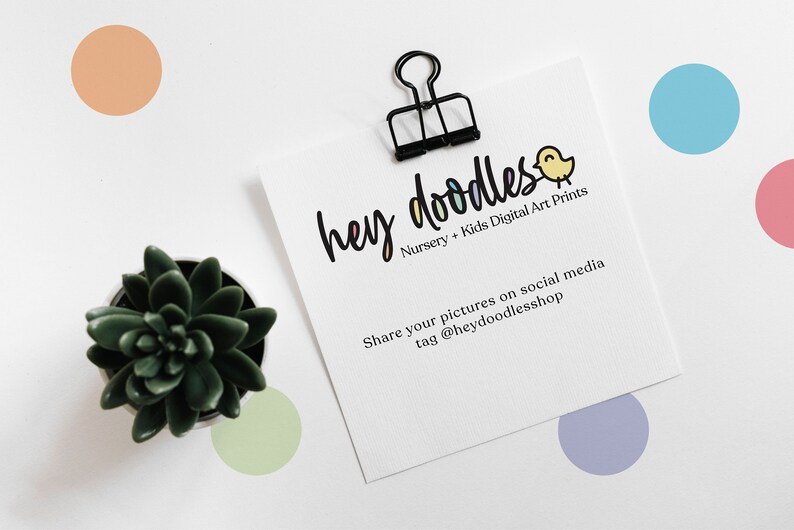 Op de afbeelding: Een witte kaart met zwarte tekst die "hey doodles Nursery + Kids Digital Art Prints Share your pictures on social media tag @heydoodlesshop" zegt. De kaart wordt vastgehouden door een zwarte paperclip. Een kleine potplant staat in de linkerbenedenhoek van de afbeelding. Er zijn kleurrijke stippen verspreid rond de kaart.