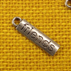 May include: Close-up of several small, rectangular metal charms with the word "friends" engraved on them. Each charm has a small loop at one end. The charms are silver-colored and rest on a textured yellow surface.