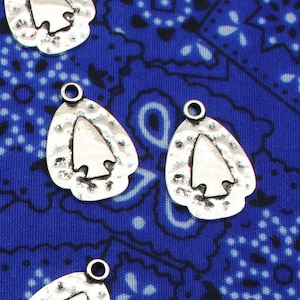 May include: A collection of silver-toned arrowhead charms with a hammered texture. The charms are displayed on a blue and white patterned fabric.