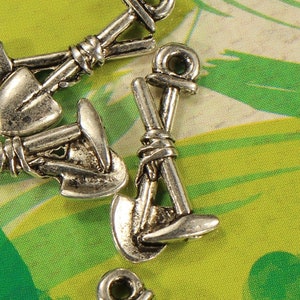 May include: Silver-toned metal charms shaped like shovels, with a heart-shaped detail on one charm. The charms are scattered on a green and white patterned surface.