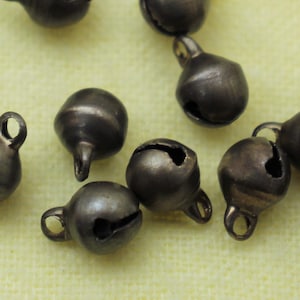 May include: A collection of small, silver-colored bells with a rounded shape and a small loop at the top. The bells are scattered on a light yellow fabric surface.