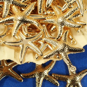 May include: A pile of gold-colored metal starfish charms in a white seashell. The charms are scattered on a blue background.