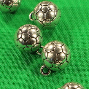 May include: Seven silver soccer ball buttons with a raised, textured design. The buttons are arranged in a diagonal line on a green surface.