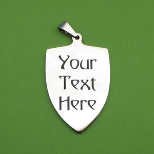 May include: A silver shield-shaped pendant with a loop at the top for attaching a chain. The pendant has the text "Your Text Here" engraved on it.