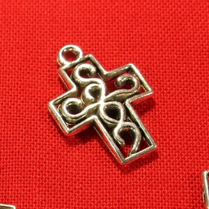 Könnte beinhalten: Silberfarbener Metallkreuz-Anhänger mit einem dekorativen, filigranen Design. Das Kreuz besteht aus einem einzigen Stück Metall mit einem filigranen Muster.