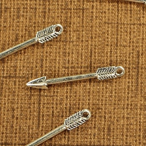 May include: Silver arrow charms with a textured design on a brown background. The charms have a pointed tip and a decorative fletching design. Each charm has a small loop at the end.