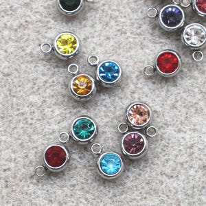 May include: A collection of small, round charms with colorful gemstones set in silver-colored metal. The gemstones are various colors, including green, blue, red, yellow, and purple. Each charm has a small loop for attaching to jewelry.