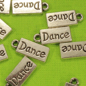 Könnte beinhalten: Eine Sammlung silberfarbener Metall-Anhänger mit dem Wort "Dance" auf jedem Anhänger. Die Anhänger sind auf einer grünen Oberfläche verstreut.