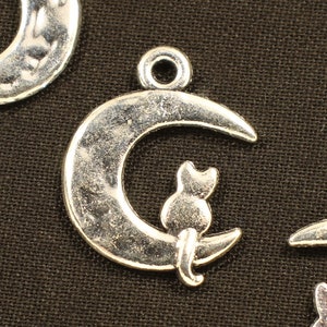 May include: Three silver charm pendants featuring a crescent moon with a cat sitting on the edge. The moon has a textured surface.