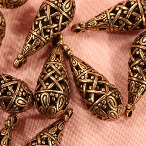 May include: A group of 13 gold-toned metal teardrop-shaped charms with intricate designs. The charms have a woven pattern and are perfect for jewelry making.