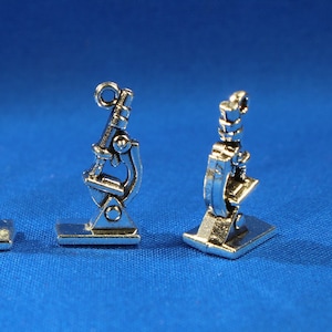 May include: Four small, silver-colored microscope charms are arranged in a row against a blue background. Each charm has a detailed design, including the eyepiece, objective lenses, and base. These charms are ideal for science-themed jewelry or craft projects.