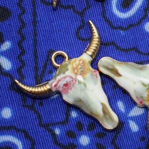 May include: Three gold-tone metal charms shaped like a bull skull with floral designs. The charms have a white background with pink, yellow, and white flowers.