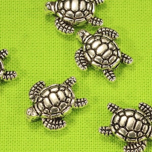 May include: Six silver turtle charms on a green background. The turtles have detailed shells and are a popular choice for jewelry making.