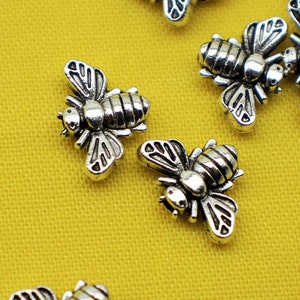 May include: A group of silver bee charms on a yellow background. The bees have detailed wings and bodies.