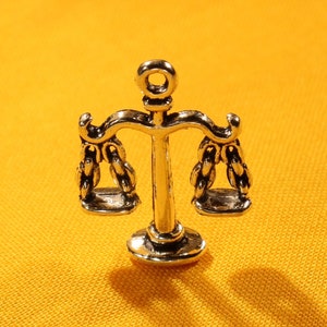 May include: A silver charm in the shape of a scale of justice. The scale is detailed with intricate designs and has a loop at the top for attaching to a necklace or bracelet.