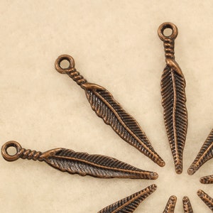 May include: Ten antique copper-toned feather charms with a loop at the top. The charms are arranged in a starburst pattern on a beige background.