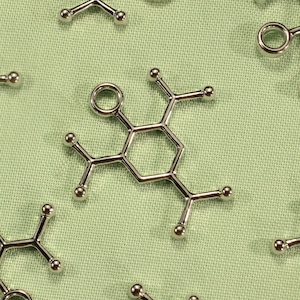 May include: A collection of silver metal charms shaped like a chemical molecule, each with a hexagonal ring and multiple small circles attached. The charms are arranged on a green fabric background.