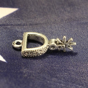 May include: Silver-toned metal charm in the shape of a horseshoe with a spur detail. The charm has an ornate design and a loop for attaching to a necklace or bracelet.