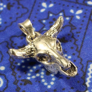 May include: Silver-toned pendant in the shape of a bull skull. The pendant is on a blue and white patterned fabric background.