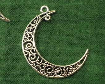 May include: Silver-toned crescent moon charms with intricate openwork scroll designs. Each charm has a small loop at the top for easy attachment. The charms are displayed on a dark green fabric background.