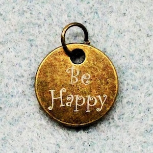 以下が含まれることがあります： ネックレスやブレスレットに取り付けるための穴が上部に開いた、丸い形の金色メタルチャーム。チャームには「Be Happy」という文字が白く刻まれています。