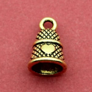 May include: A small, gold-colored bell-shaped charm with a heart design. The charm has a loop at the top for attaching to jewelry or other crafts. The bell has a textured pattern and is set against a red background.