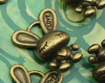 Pode incluir: Encantos de coelho de cor bronze com a palavra "LOVE" nas orelhas. Os encantos têm uma aparência vintage e estão espalhados sobre um fundo verde padronizado. Ideais para fazer joias ou projetos de artesanato.