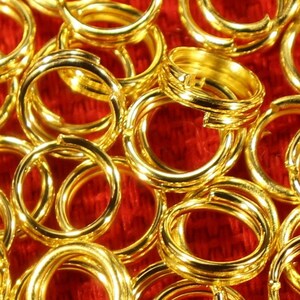 May include: A close-up image of a large quantity of gold jump rings. The rings are scattered on a red fabric surface.