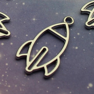May include: Silver metal rocket ship charms with a simple outline design. These charms are perfect for adding a space-themed touch to jewelry or crafts.