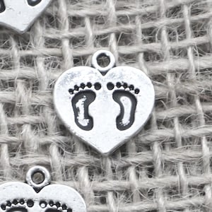 May include: Silver-toned heart-shaped charms with baby footprints. Each charm has a small loop at the top for attaching to jewelry or other crafts. The charms are made of a metal alloy and have a slightly distressed finish.