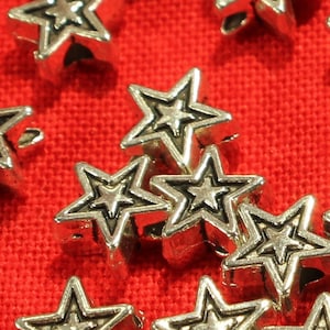 May include: A close-up of a group of silver star-shaped beads on a red fabric background. The beads have a raised design and are perfect for jewelry making or crafting.