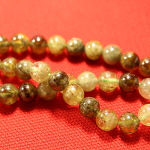 May include: A strand of green and brown gemstone beads on a red fabric background. The beads are round and have a natural, unpolished look.