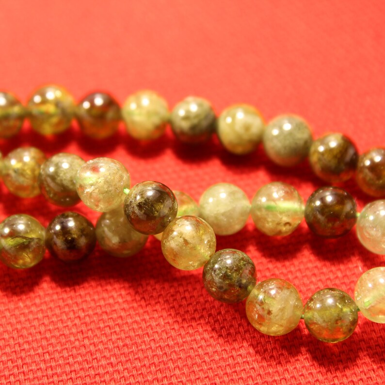 May include: A strand of green and brown gemstone beads on a red fabric background. The beads are round and have a natural, unpolished look.