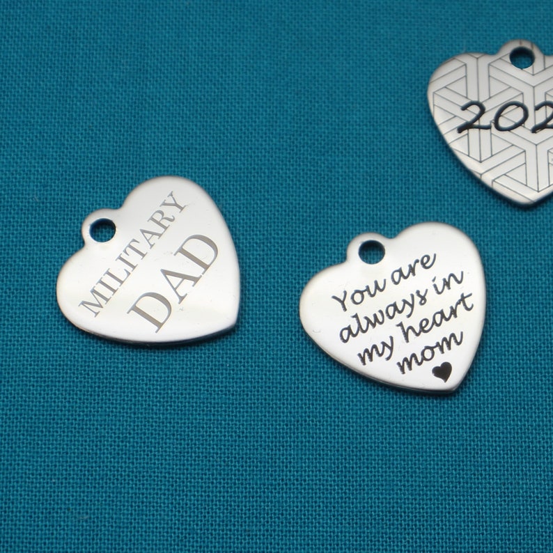 May include: Three silver heart-shaped charms with engraved messages. The first charm reads "Military Dad", the second charm reads "You are always in my heart mom", and the third charm has a geometric pattern and the number "2021" engraved on it.
