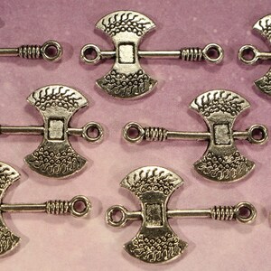 May include: Silver tone axe-shaped metal charms with a decorative design. Each charm has a loop on either side for attaching to a chain or bracelet.
