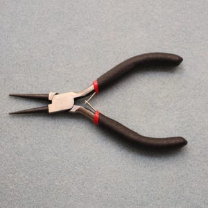 May include: Round nose pliers with black handles and red accents. The pliers have a silver metal head and long, pointed tips. These pliers are used for jewellery making, wire wrapping, and other craft projects.