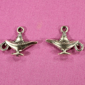 May include: Two silver-toned genie lamp charms with intricate details. The lamps have a rounded base and a spout with a loop handle. The charms are on a pink fabric background.